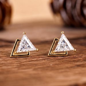 May include: A pair of gold stud earrings featuring a triangular clear gemstone set within a geometric gold frame. Each earring consists of a central triangle-cut stone positioned next to an open triangular gold metal outline, creating a layered look.