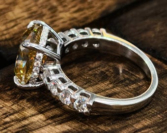 May include: A silver ring with a large, oval-cut yellow gemstone as the centerpiece. The band is adorned with smaller, clear gemstones. The ring is set against a dark wooden surface, highlighting its sparkle.