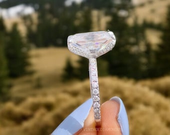 Può includere: Un anello di fidanzamento in argento con un grande diamante taglio smeraldo trasparente. L'anello ha una montatura a pavé con diamanti più piccoli sulla fascia. L'anello è tenuto da una mano su uno sfondo sfocato di montagne.