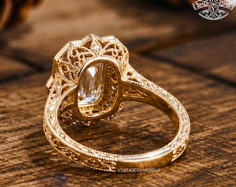 May include: Close-up of a gold engagement ring with an emerald-cut diamond. The ring features intricate filigree detailing and a vintage design. The band is engraved with a delicate pattern. The text "VINTAGEDIAMONDS" is visible on the ring.