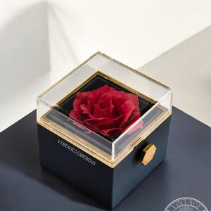 May include: A single red rose preserved in a clear glass box with a gold trim. The box is sitting on a dark blue surface with the text "VINTAGEDIAMONDUS" printed on the side. The box has a gold hexagonal knob on the top.