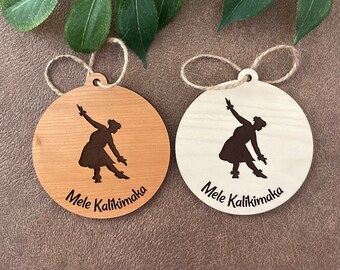 Puede incluir: Dos adornos redondos de madera con una silueta de bailarina y el texto "Mele Kalikimaka". Uno es marrón claro, el otro blanco. Ambos tienen un lazo de cuerda para colgar.