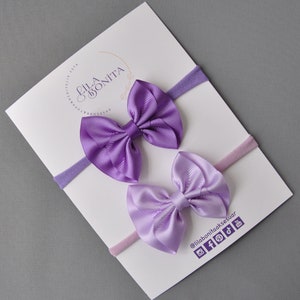May include: Two purple and lavender satin ribbon bows attached to nude and pink elastic headbands. The headbands are displayed on a white card with the text "Lila Bonita" in a gold script font. The card also includes social media handles for the brand.