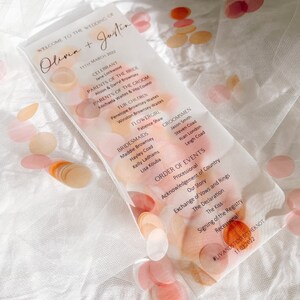 Puede incluir: Orden del día de la ceremonia de boda impreso en una hoja de acrílico transparente con un fondo de acuarela de círculos rosas y naranjas. El texto incluye los nombres Olivia y Justin, la fecha 11 de marzo de 2022 y el orden del día de la ceremonia de boda.