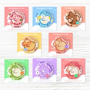 Könnte beinhalten: Eine Sammlung bunter runder Buttons mit niedlichen Cartoon-Tieren und verschiedenen Getränkearomen. Jeder Button trägt den Text "honeypeach.studio" und den Getränkegeschmack, wie "Strawberry Matcha" und "Blueberry Lemonade".