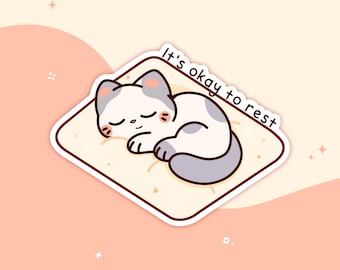 Könnte beinhalten: Ein Aufkleber mit einer Cartoon-Katze, die auf einem hellgelben Kissen schläft. Die Katze ist weiß mit grauen Markierungen und geschlossenen Augen. Der Aufkleber hat den Text "It's okay to rest" in handschriftlicher Form.