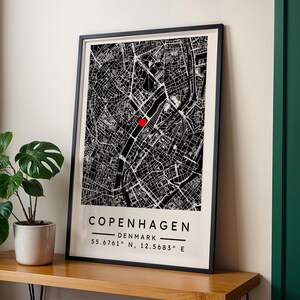 May include: Framed black and white map of Copenhagen, Denmark, with a red heart marking a location. The city name and coordinates are printed below the map. The framed print is displayed on a wooden surface.