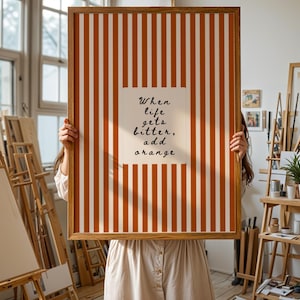 May include: A framed artwork with a vertical orange and white striped background. A white square in the center features the quote, "When life gets bitter, add orange." The frame is a warm brown color. The artwork is held by a person.
