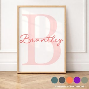 May include: A rectangular wall art print in a light wood frame featuring a large, pale pink capital letter B as the background. Overlaid across the center of the letter is the name Brantley written in a cursive, dark pink script font.