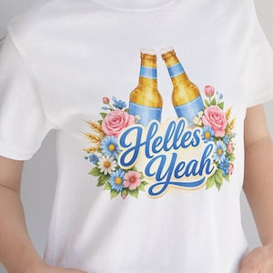 May include: A white t-shirt featuring a graphic of two clinking beer bottles surrounded by pink roses, blue daisies, and wheat stalks. The text "Helles Yeah" is printed in a blue script font across the center of the floral arrangement.