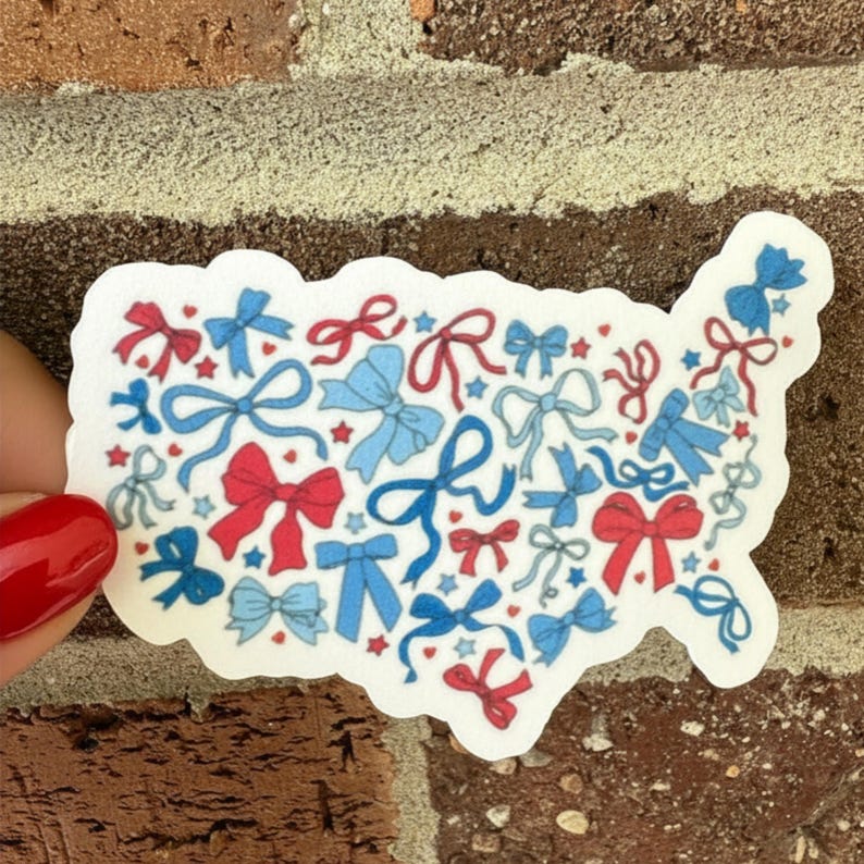 May include: A white sticker in the shape of the United States, filled with red and blue bows and stars. The sticker is held by a hand with a red painted fingernail, against a brick background.