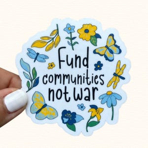 May include: A white sticker with the text "Fund communities not war" in black. The sticker is decorated with blue and yellow butterflies, flowers, and leaves. The sticker is held by a person.
