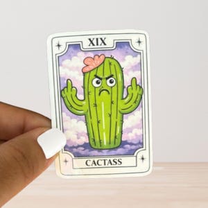 May include: A rectangular sticker designed like a tarot card featuring a green cartoon cactus with a pink flower on top, an angry facial expression, and arms raised giving the middle finger. The text XIX and CACTASS are printed on the card.