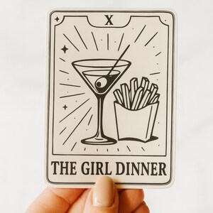 May include: A rectangular sticker with a black and white illustration of a martini glass with an olive, a side of fries, and the text "THE GIRL DINNER". The sticker has a tarot card design with the number "X" at the top.