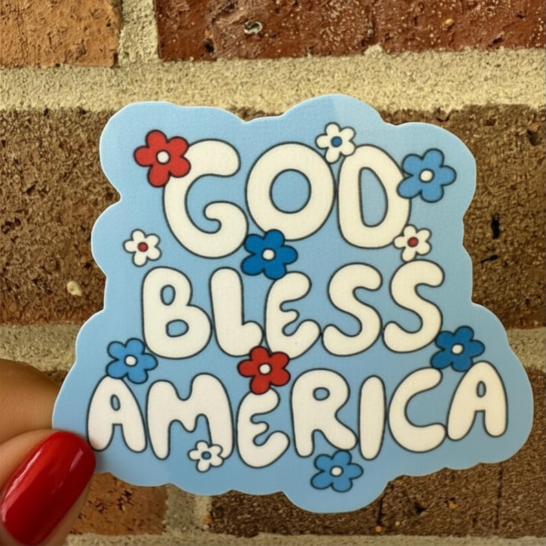 May include: A light blue sticker with the words "GOD BLESS AMERICA" in white, decorated with red, white, and blue flowers. The sticker is held against a brick wall.