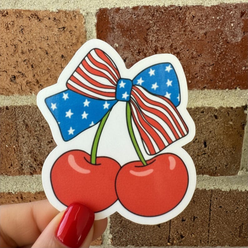 May include: A sticker featuring a pair of red cherries with green stems and a patriotic bow. The bow has blue sections with white stars and red and white stripes, resembling the American flag.