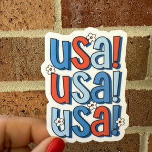 May include: A white sticker with the word "USA!" repeated three times in red and blue, with small white flowers. The sticker is held by a hand with red nail polish, against a brick background.