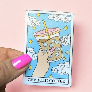 May include: A tarot card illustration featuring an iced coffee drink in a clear cup with ice cubes, a pink lid, and a yellow straw. The card has a light blue background with white clouds and yellow stars. The text "THE ICED COFFEE" is at the bottom.
