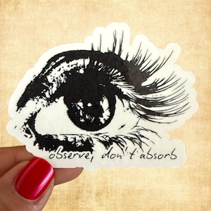 May include: A die-cut sticker featuring a high-contrast black and white illustration of a human eye with long, detailed eyelashes. Below the eye, the text "observe, don't absorb" is written in a simple, handwritten black script font.