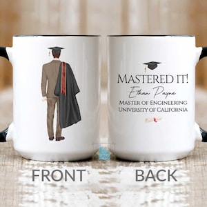 May include: White ceramic mug with a black handle and rim. One side features a graduate in a suit and graduation gown. The other side says "MASTERED IT!" with a name, degree, and university information. A graduation cap and diploma are also shown.