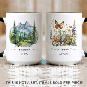 Puede incluir: Dos tazas de cerámica blanca con asas y bordes negros. La taza izquierda presenta una escena de acuarela de montañas y bosque de pinos con el texto "nonno est 2026". La taza derecha muestra un ramo de flores silvestres y una mariposa con el texto "nonna est 2026".