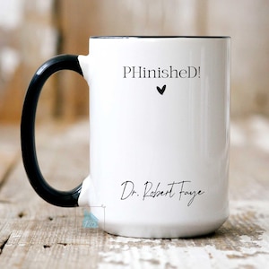 May include: White ceramic mug with a black handle and rim. The mug features the text "PHinished!" above a small black heart, and the name "Dr. Robert Faye" in a script font.