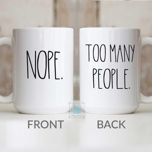 May include: Two white ceramic mugs with black text. One mug reads "NOPE." on the front. The other mug reads "TOO MANY PEOPLE." on the back. Both mugs have a white handle and a simple, modern design.