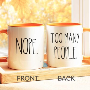 May include: Two white ceramic mugs with orange interiors and handles. One mug reads "NOPE." in black text, and the other reads "TOO MANY PEOPLE." The mugs sit on a wooden tray.