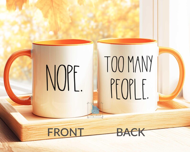 May include: Two white ceramic mugs with orange interiors and handles. One mug reads "NOPE." in black text, and the other reads "TOO MANY PEOPLE." The mugs sit on a wooden tray.