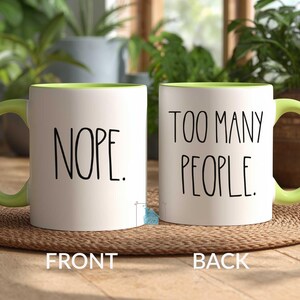 May include: Two white ceramic mugs with green handles and interior rims. One mug reads "NOPE." in black text, and the other reads "TOO MANY PEOPLE." The mugs are on a woven mat, with plants in the background.