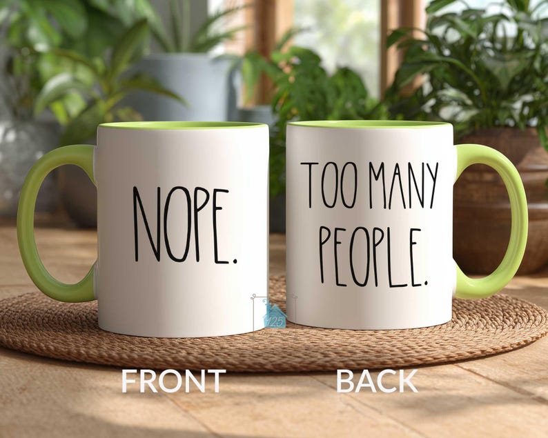 May include: Two white ceramic mugs with green handles and interior rims. One mug reads "NOPE." in black text, and the other reads "TOO MANY PEOPLE." The mugs are on a woven mat, with plants in the background.