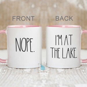 May include: White ceramic coffee mug with a pink rim and handle. The front of the mug says "NOPE." The back of the mug says "I'M AT THE LAKE."