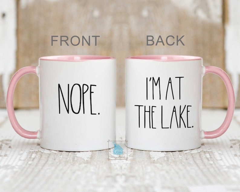 May include: White ceramic coffee mug with a pink rim and handle. The front of the mug says "NOPE." The back of the mug says "I'M AT THE LAKE."