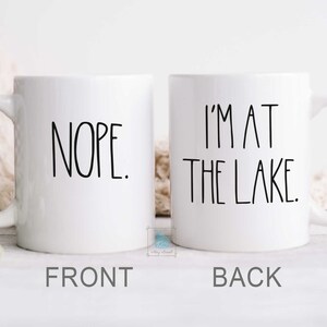 May include: White ceramic coffee mug with black text. The front of the mug says "NOPE." The back of the mug says "I'M AT THE LAKE."
