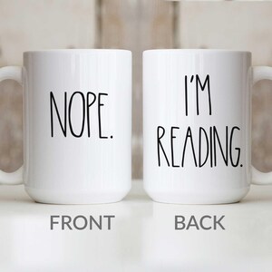 May include: White ceramic coffee mug with black text. The front of the mug says "NOPE." The back of the mug says "I'M READING."