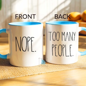 May include: White ceramic mug with a light blue interior, handle, and rim. The front of the mug reads "NOPE." in black text, and the back reads "TOO MANY PEOPLE." A humorous coffee mug.