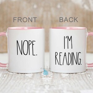 May include: White ceramic coffee mug with a pink rim. The front of the mug says "NOPE." The back of the mug says "I'M READING."