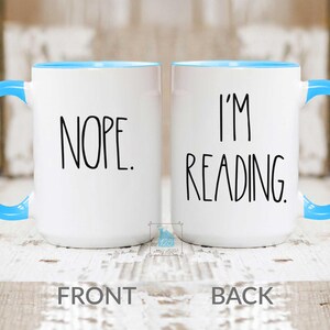 May include: White ceramic mug with a blue rim. The front of the mug says "NOPE." The back of the mug says "I'M READING."