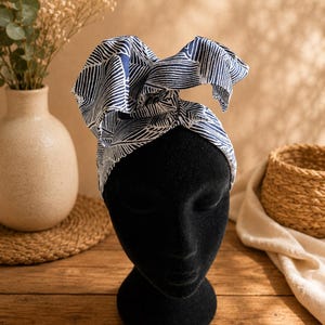 May include: A fabric headwrap featuring a dark blue and white abstract leaf pattern. The headwrap is tied in a large, prominent bow at the top and is displayed on a smooth, matte black mannequin head, showcasing the intricate linear design.