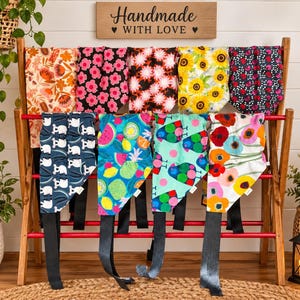 May include: A wooden rack displaying nine fabric bandanas with various patterns, including floral, fruit, and animal prints. Each bandana features black fabric ties at the bottom. A wooden sign above the rack reads Handmade WITH LOVE.