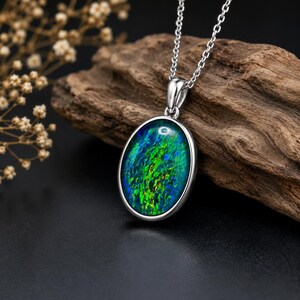 May include: Silver pendant necklace featuring an oval opal gemstone with vibrant green and blue hues. The gemstone is set in a silver bezel and hangs from a delicate silver chain. The necklace is displayed on a piece of driftwood against a dark background.