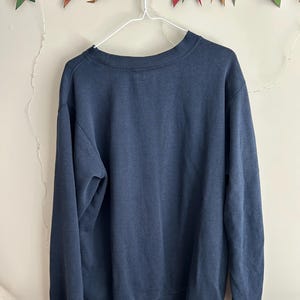 May include: A solid navy blue crewneck sweatshirt with long sleeves and ribbed cuffs. The garment is displayed on a white wire hanger, featuring a simple, plain design with no visible graphics, patterns, or text on the front of the fabric.