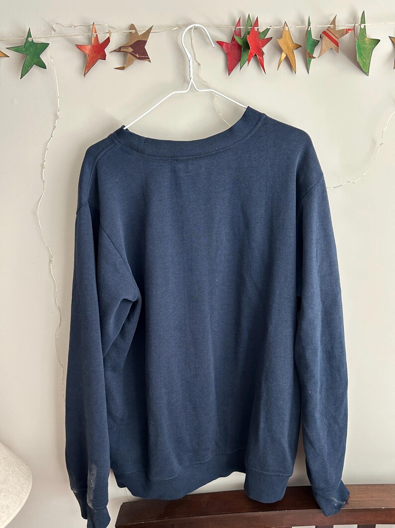May include: A solid navy blue crewneck sweatshirt with long sleeves and ribbed cuffs. The garment is displayed on a white wire hanger, featuring a simple, plain design with no visible graphics, patterns, or text on the front of the fabric.