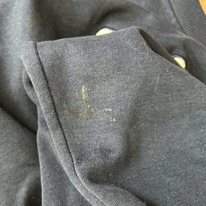 May include: A dark navy blue sweatshirt featuring a ribbed collar and a visible, worn-in Nike swoosh logo on the chest. The fabric shows signs of wear, including light pilling and a small, faded, yellowish stain near the embroidered logo area.
