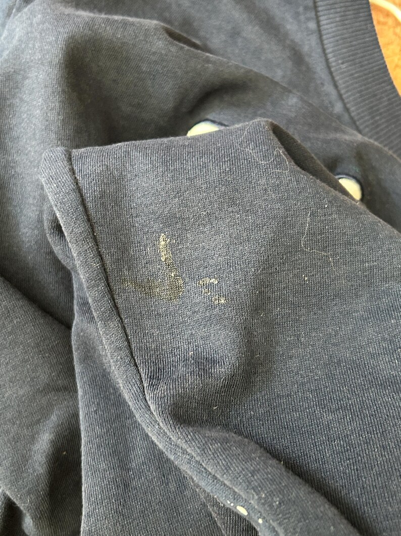 May include: A dark navy blue sweatshirt featuring a ribbed collar and a visible, worn-in Nike swoosh logo on the chest. The fabric shows signs of wear, including light pilling and a small, faded, yellowish stain near the embroidered logo area.