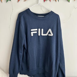 May include: A navy blue long-sleeve sweatshirt featuring a prominent white FILA logo appliqued across the chest. The garment has a ribbed crew neckline, ribbed cuffs, and a ribbed hem, constructed from a solid-coloured fabric with a soft texture.