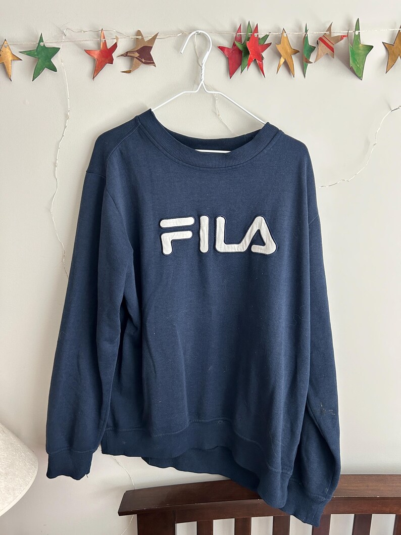 May include: A navy blue long-sleeve sweatshirt featuring a prominent white FILA logo appliqued across the chest. The garment has a ribbed crew neckline, ribbed cuffs, and a ribbed hem, constructed from a solid-coloured fabric with a soft texture.