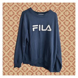 May include: A navy blue long-sleeve sweatshirt featuring a crew neckline and ribbed cuffs. The front of the garment displays the brand name FILA in large, raised white block letters across the chest, constructed with a textured, padded finish.