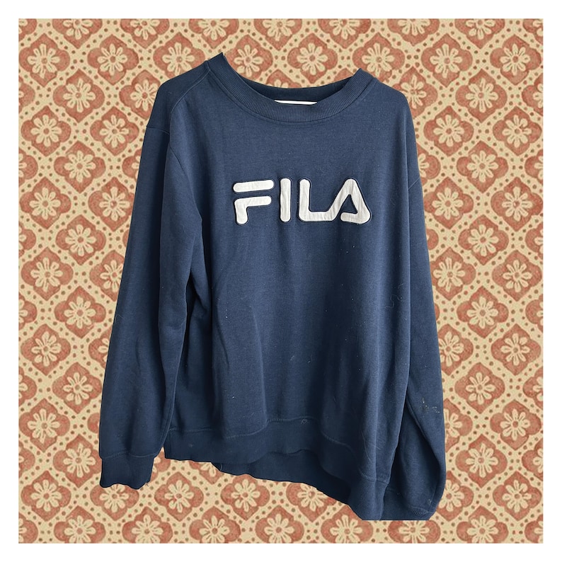 May include: A navy blue long-sleeve sweatshirt featuring a crew neckline and ribbed cuffs. The front of the garment displays the brand name FILA in large, raised white block letters across the chest, constructed with a textured, padded finish.
