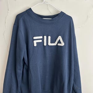 May include: A navy blue long-sleeve sweatshirt featuring a crew neckline and ribbed cuffs. The front of the garment displays the word FILA in large, raised, white block letters with a dark blue outline, centred on the chest area.